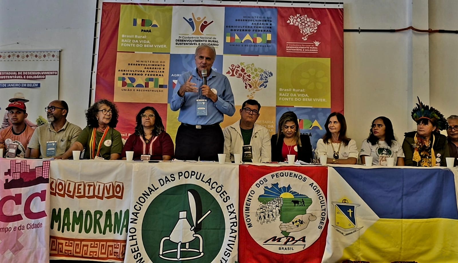 Imagem mostra mesa com dez pessoas sentadas e um homem branco, alto, de camisa azul e calça preta ao centro com microfone falando 