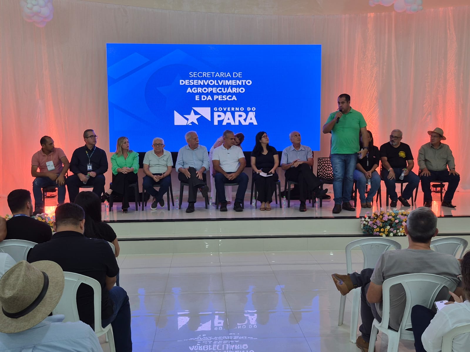 imagem mostra 11 pessoas sentadas e uma de pé em cadeiras, na frente de um auditório