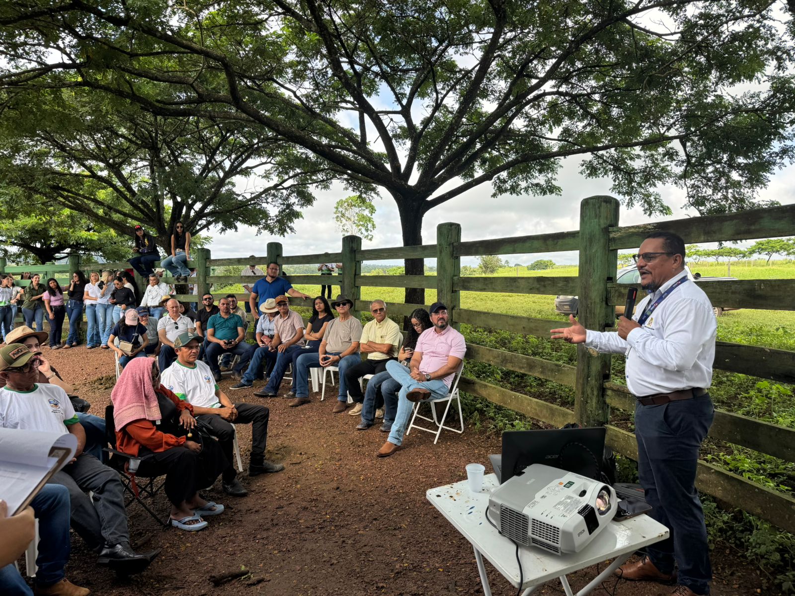 Imagem mostra homem negro, de camisa branca e calça escura, de pé, falando para grupo sentado em cadeiras distribuídas em área aberta sob árvores