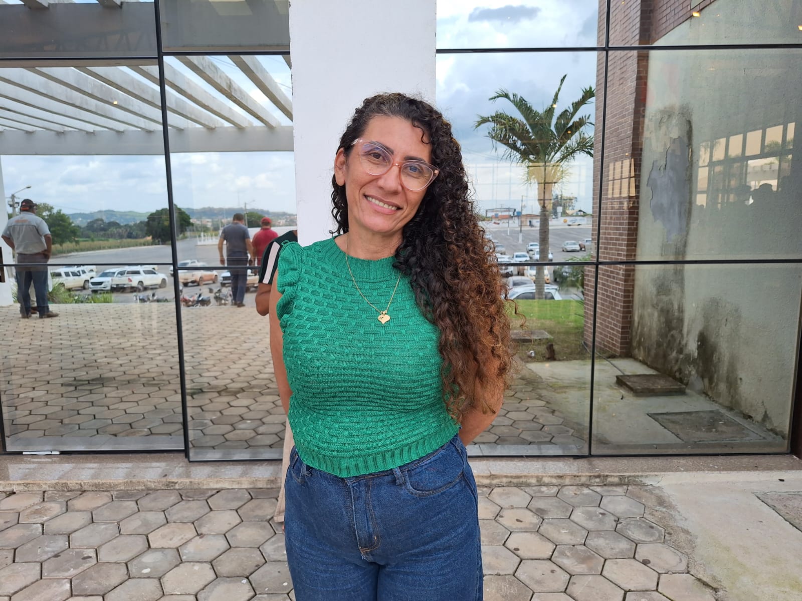 Agricultora, de camisa verde e calça e jeans, cm braços para trás, de pé e sorridente