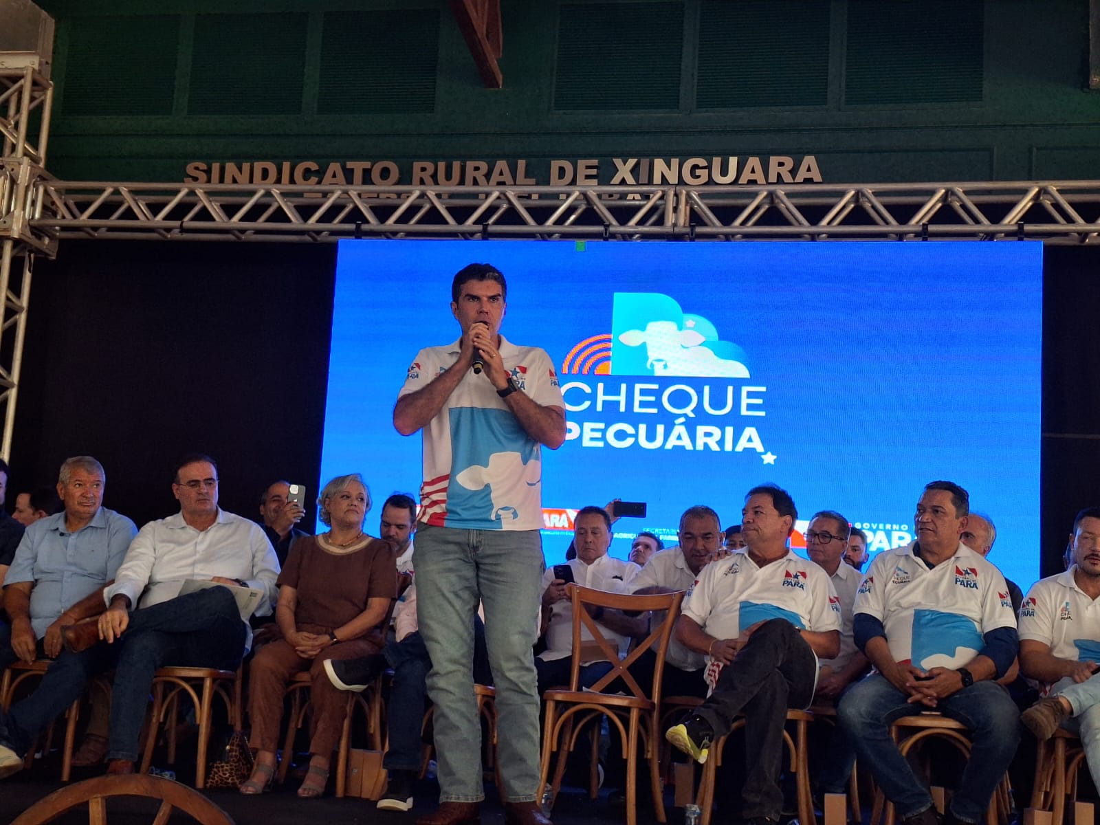 Palco mostra governador falando, de pé ao microfone e várias outras pessoas sentadas na lateral e atrás