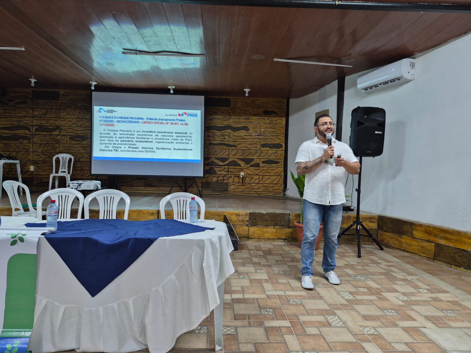 Homem branco, de camisa clara, estatura mediana, meia idade, falando ao microfone ao lado de uma mesa e com projeção de imagens atrás dele