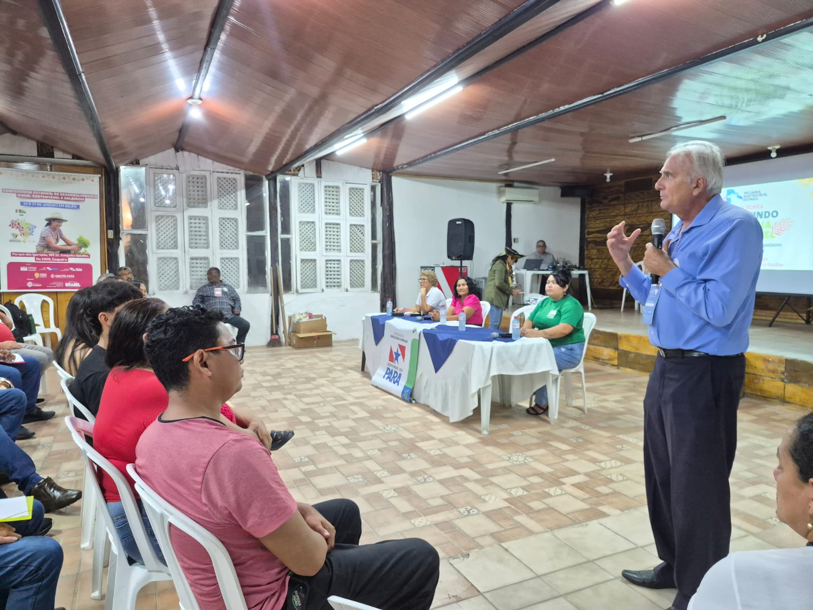 Imagem mostra homem branco,, alto, de camisa azul clara e calça escura, falando ao microfone para público em sala, durante palestra técnica