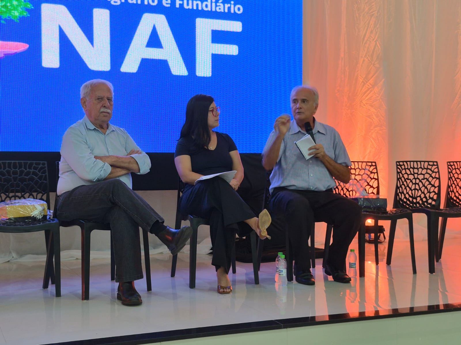 Imagem mostra três pessoas, dois homens e uma mulher, sentados à frente de auditório, um deles com microfone a falar para o público