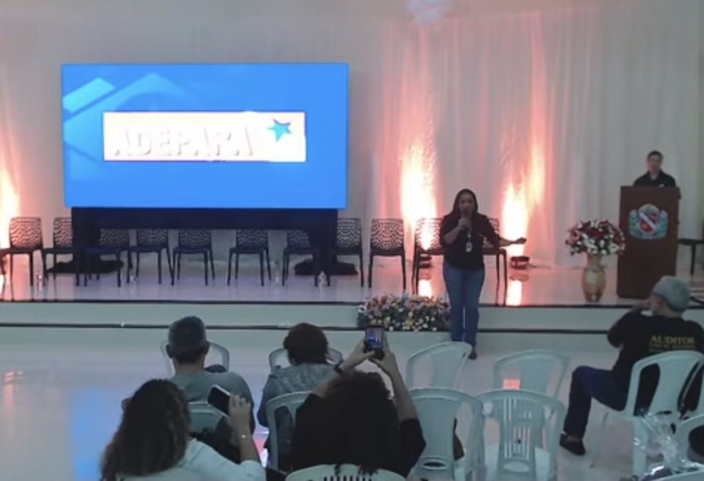 Imagem mostra mulher ao microfone à frente de auditório, outra mulher ao fundo  e um telão na lateral de ambas