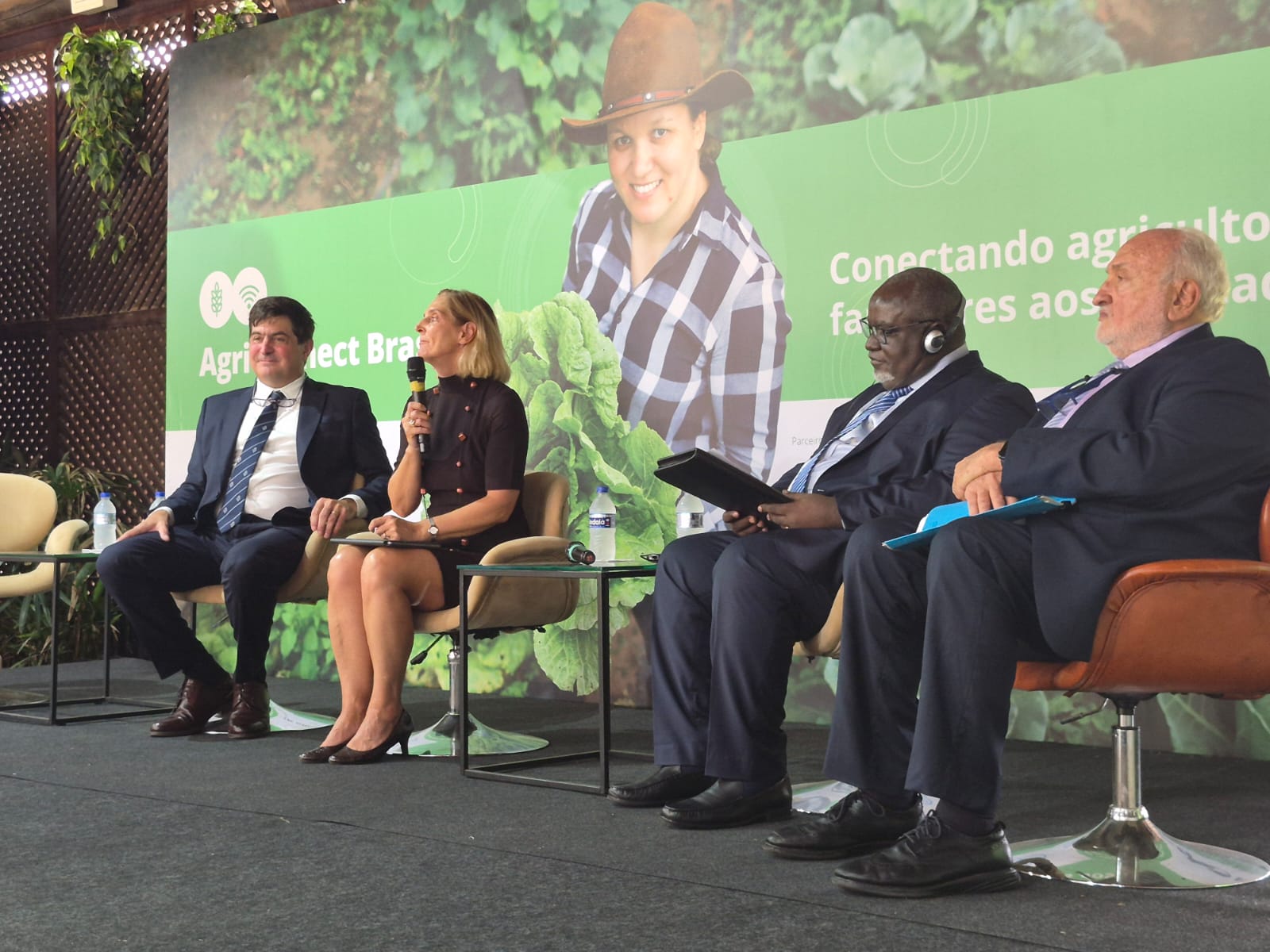 Imagem mostra mulher branca com microfone na mão falando ao público com dois três homens ao lado, de etinias diversas