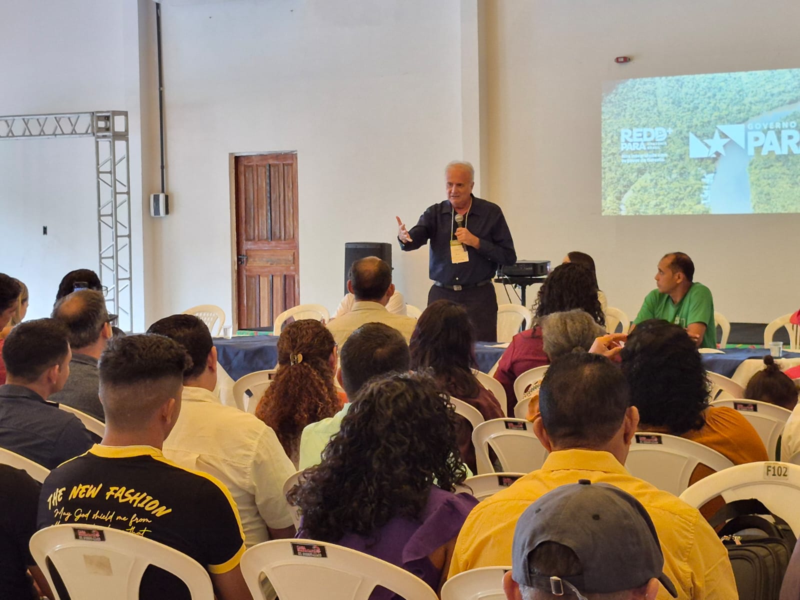 Imagem mostra homem branco de camisa azul, de pé, falando para ppublico em auditório