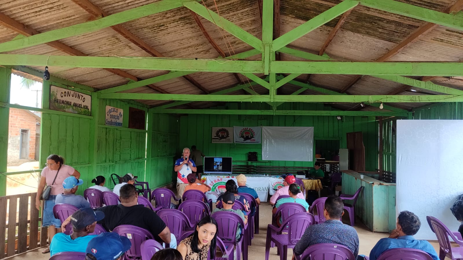 Imagemmostra púbico em barracão de madeira assistindo apresentação de homem ao microfone