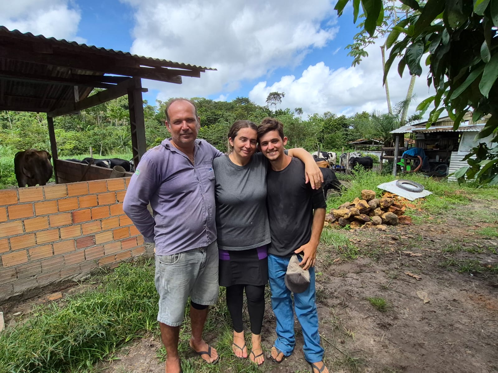 Imagem mostra casal de idade média e filho jovem lado a lado em área verde com vacas atrás