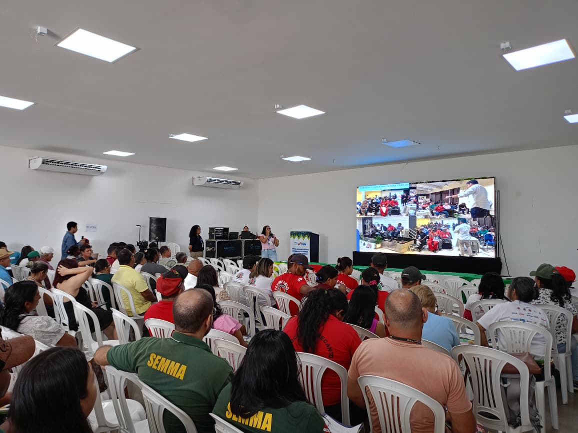 Imagem mostra auditório com pessoas sentadas assistindo atividades em um telão e uma pessoa ao microfone 