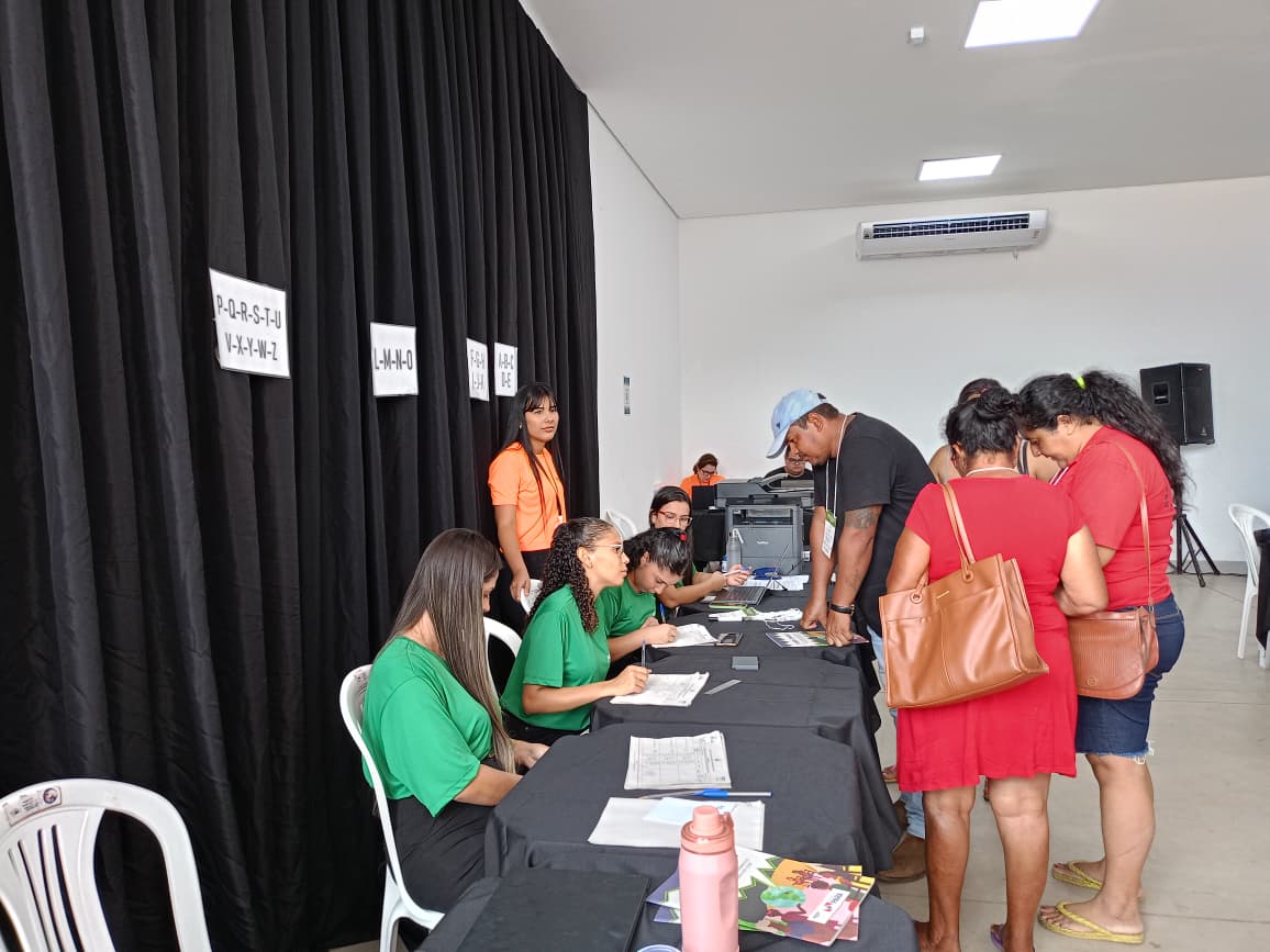 Imagem mostra pessoas de pé à frente de uma mesa, com outras sentadas em troca de informações e assinaturas